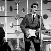 Buddy Holly playing guitar on stage, wearing his signature glasses and smiling.