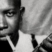 A black-and-white portrait of Robert Johnson, the legendary blues musician, captured in a formal pose, wearing a suit and looking directly at the camera.
