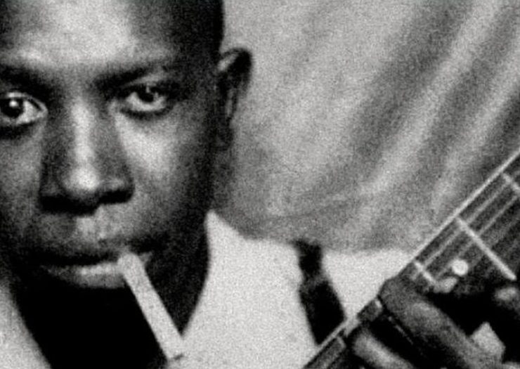 A black-and-white portrait of Robert Johnson, the legendary blues musician, captured in a formal pose, wearing a suit and looking directly at the camera.